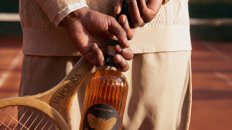 Person holding a bottle of Vapoura Chardonnay with a tennis racket on a tennis court.